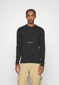 Felpa nera a maglia con maniche lunghe e polsini a costine, con logo bianco "Calvin Klein Jeans" al centro davanti.