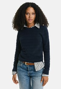 Mörkblå stickad tröja med ribbat mönster, med randig skjortkrage och manschetter. Kombinerad med ljusblå jeans och ett brunt bälte.