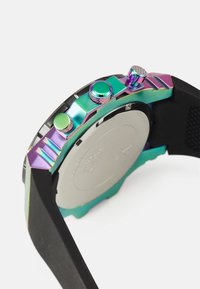 Armbåndsur med en flerfarget metallkasse som har en grønn og lilla gradient, teksturerte knapper og et sort silikonearmbånd.