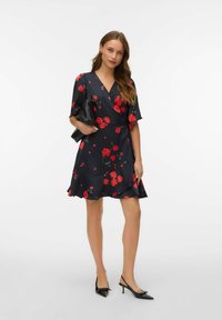 Robe portefeuille à motifs floraux dans un bleu marine foncé avec des roses rouges, manches courtes à volants, ourlet avec volants, accompagnée d'une pochette noire et de talons à bout pointu.