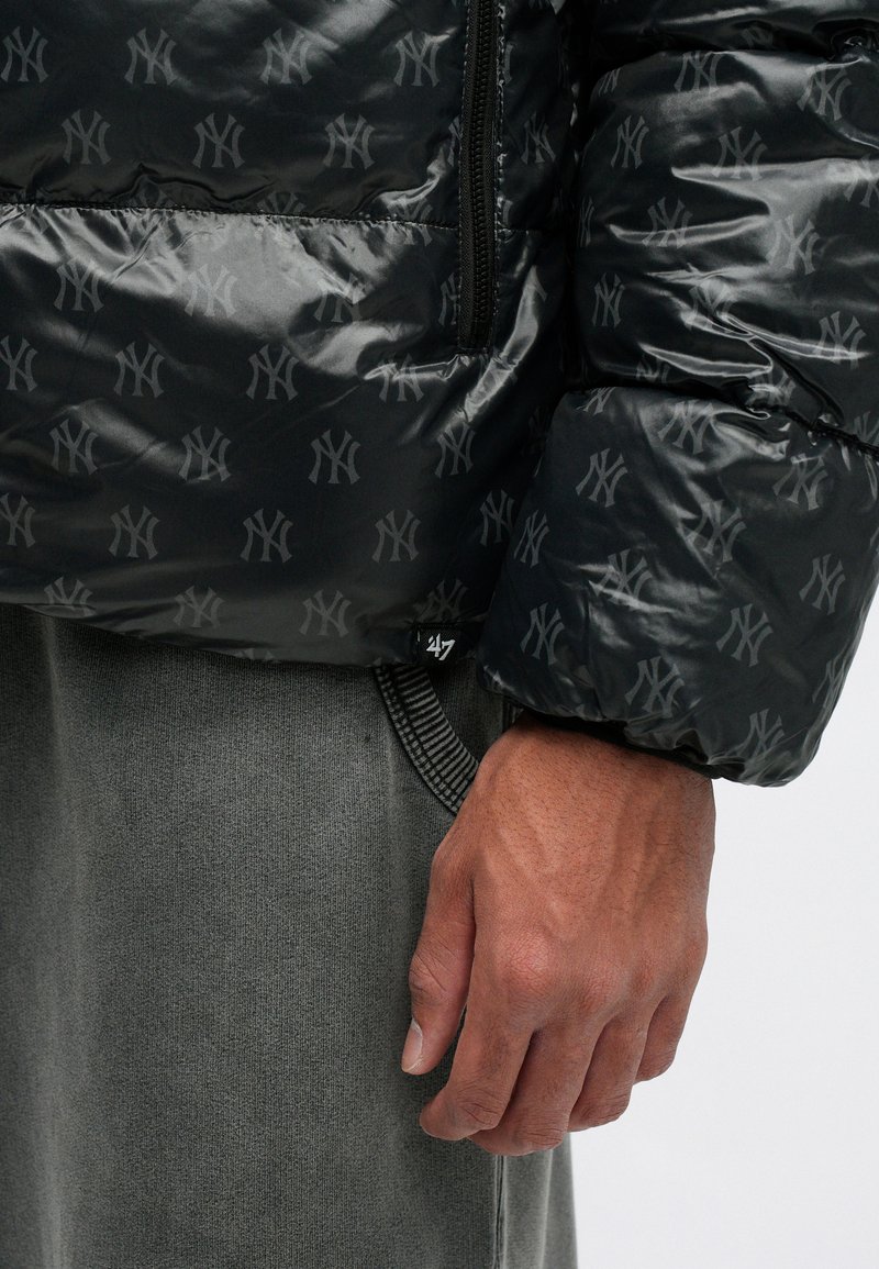 Schwarze Daunenjacke mit tonalem NY-Logo-Muster, glänzender Oberfläche, Reißverschluss und gerippten Bündchen. Grauer Pullover darunter sichtbar.