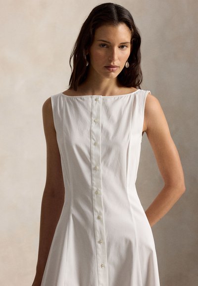Woman with dark hair wearing sleeveless white button-down dress and white drop earrings, standing against a neutral background.