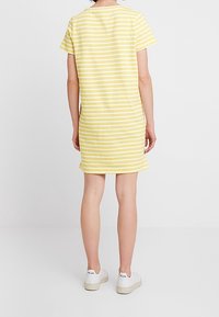 Nainen, joka on pukeutunut lyhythihnaiseen, polvimittaisiin kelta-valkoisiin raidallisiin mekkoihin ja valkoisiin lenkkareihin, seisoo selkä kameraan päin tasaisella taustalla.