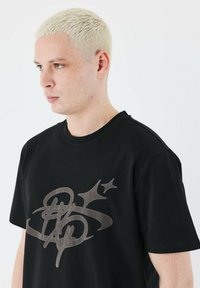 Camiseta negra de manga corta con un gran diseño gráfico gris que presenta curvas y estrellas en el frente. Hecha de material de algodón suave.
