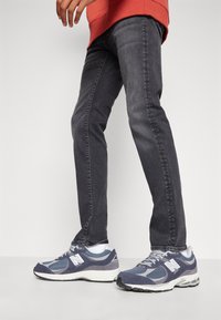 Moški sivi denim kavbojke v kombinaciji z mornarsko modrimi New Balance supergami, ki vključujejo mrežaste in semiš materiale ter bele detajle.