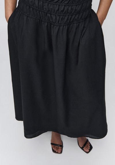 Μαύρη μίντι φούστα με ραμμένη ελαστική μέση και πλαϊνές τσέπες, φορεμένη με μαύρα σανδάλια με τετράγωνη μύτη.
