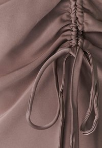 Sasietais audums mīksta taupe krāsā ar saitēm, demonstrējot gludu tekstūru un elegantu dizainu, ar vieglām savilkumiem gar šuvi.