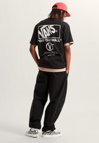 Zwart katoenen T-shirt met wit grafisch logo op de achterkant, gecombineerd met losse zwarte broek en slip-on schoenen met zebraprint, rode pet op het hoofd.