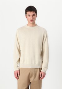 Ανοιχτό μπεζ πουλόβερ με μακριά μανίκια και ριμπ γιακά. Μαλακή υφή και εφαρμοστά μανίκια. Συνδυάζεται με καφετί παντελόνι.