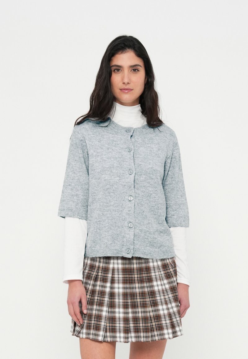 Ανοιχτό γκρι πλεκτή ζακέτα με κοντά μανίκια, στρογγυλή λαιμόκοψη και έξι κουμπιά. Συνδυασμένη με καφέ και λευκή φούστα καρό.