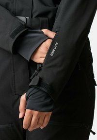 Zwarte waterdichte jas met een gladde textuur, voorzien van een ritssluiting en klittenbandsluiting op de mouw. Het label zegt "STYX 8000."