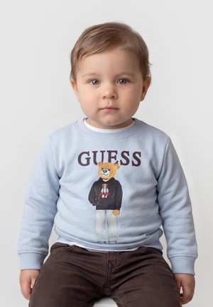 Lille barn med lysbrunt hår iført en lyseblå sweatshirt med et tøjdyr og ordet "GUESS,", siddende mod en ensfarvet baggrund.