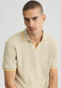 Helles beigefarbenes Poloshirt mit strukturiertem Kragen und gelben Akzenten. Verfügt über einen V-Ausschnitt und kurze Ärmel, mit dezentem Logo-Branding auf der Brust.