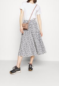 Jupe midi à motifs noir et blanc avec un design plissé, associée à un t-shirt blanc et des chaussures de sport noires. Un petit sac bandoulière marron est porté.