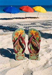 Infradito con motivi floreali e leopardati stanno in piedi nella sabbia bianca con ombrelloni da spiaggia blu, rossi e gialli e l'oceano sullo sfondo.