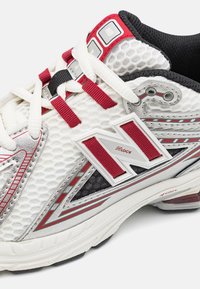 Chaussure de sport blanche avec des accents rouges et argentés, tige en maille texturée, col rembourré et logo 'N' bien en vue. Présente un système de laçage.