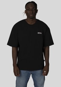 Černé oversized tričko z bavlny s bílým nápisem "CRYSTAL" na levé části hrudi, doplněné modrými džíny.