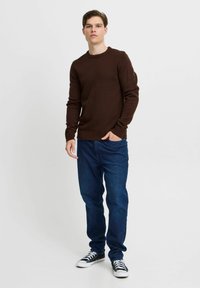 Brauner Strickpullover mit gerippten Bündchen und Saum, kombiniert mit dunkelblauen Jeans und schwarzen Sneakers mit weißen Akzenten. Das Model steht lässig.