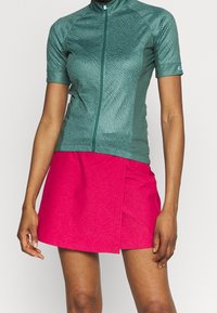 Kortärmad cykeltröja i teal med vågmönster, ihopparad med en rosa kjol med en slits fram. Tillverkad av lätt, texturerat material.