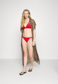 Biquíni vermelho com um top em triângulo e parte de baixo correspondente, complementado com um vestido de banho transparente de padrão leopardo em marrom, e chinelos pretos.