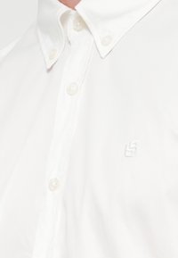 Λευκό πουκάμισο με κουμπιά από απαλό βαμβακερό ύφασμα, με κλασικό γιακά και διακριτικό κεντημένο λογότυπο στο αριστερό στήθος.