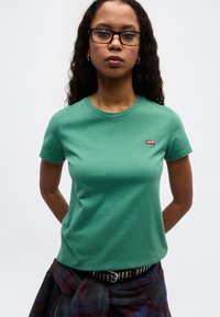 Νεαρή γυναίκα με μακριά σγουρά μαλλιά και γυαλιά, που φοράει πράσινο μπλουζάκι Levi's και καρό πουκάμισο δεμένο γύρω από τη μέση της, με φόντο λευκό.