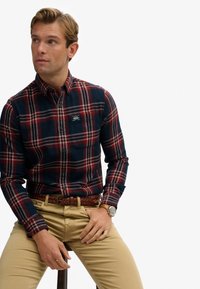 Chemise à carreaux bleu marine et rouge avec une poche, portée avec un pantalon khaki et une ceinture tressée marron. Comprend une montre avec un cadran sombre.