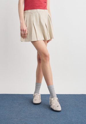 Άτομο που φοράει μπεζ πλισέ φούστα, κόκκινο τοπ, γκρι κάλτσες και λευκά αθλητικά παπούτσια, στέκεται σε μπλε χαλί μπροστά σε λευκό, απλό τοίχο.