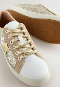 Baltie sporta apavi ar mirdzošiem zelta akcentiem, bēša zamšādas pārklājumiem un metāliskiem šņorēm. Iezīmē teksturētu gumijas zoli ar rakstainu saķeri.