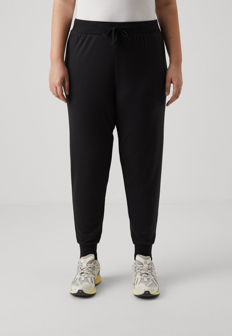 Joggers noirs fuselés avec taille à cordon, poignets côtelés et texture lisse, associés à des baskets de sport légères.