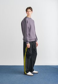 Lila sweatshirt med svarta ränder, kombinerad med svarta byxor med gula sidostrimmor. Vita sneakers och en svart keps bärs.