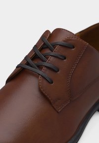 Καφέ δερμάτινο επίσημο παπούτσι με μαύρα κορδόνια, με λεία υφή και λεπτομερείς ραφές κοντά στις τρύπες των κορδονιών και στα πλαϊνά πάνελ.