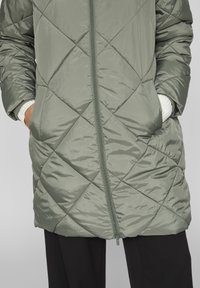 Gepolsterter, gesteppter Mantel in gedämpftem Grün. Verfügt über einen vollständigen Reißverschluss, zwei seitliche Taschen und gerippte weiße Bündchen, die aus den Ärmeln herausblicken.