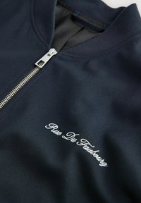 Gros plan sur une veste bleu marine avec une fermeture éclair argentée et un texte brodé blanc "Rue Du Faubourg" sur le tissu près du col.