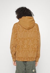 Persona con cabello rizado que lleva un sudadera con capucha de patrón marrón y pantalones cargo verdes, de espaldas frente a un fondo sencillo.