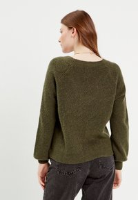 Pull en tricot vert avec un col large, une texture côtelée et de longues manches. L'ourlet est légèrement plus long à l'arrière, associé à un pantalon sombre.