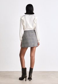 Witte blouse met gerimpelde schouders, gecombineerd met een grijze geruite mini rokje. Zwarte hakken enkellaarsjes maken de outfit compleet. Minimalistische achtergrond.