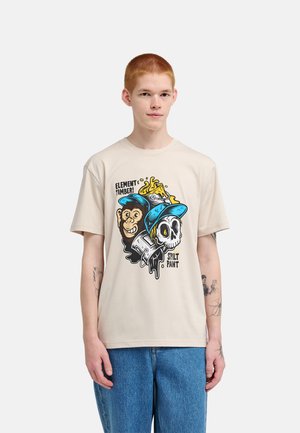 Giovane che indossa una t-shirt beige con grafica colorata di una scimmia e un teschio con tappi di vernice e testo "Element Timber! Spilt Paint."