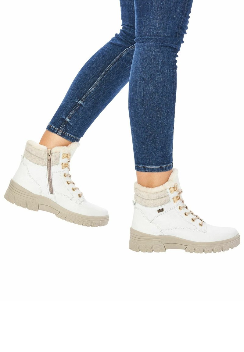 Persoon die een blauwe skinny jeans en witte veterwinterlaarzen met beige zolen en fleece voering draagt, met één voet iets omhoog geheven.