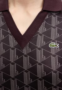 Gros plan d'une personne portant un t-shirt à col en V sombre à motifs avec un petit logo vert de crocodile sur le côté droit.