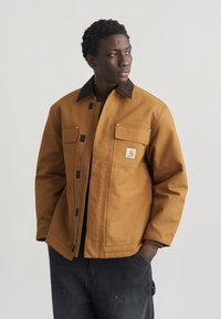 Άνδρας που φοράει καφέ μπουφάν Carhartt με τσέπες με καπάκι και μαύρο παντελόνι, κοιτάζοντας πλάγια με το ένα χέρι στην τσέπη, σε απλό φόντο.