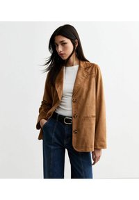 Bruine faux suede blazer met notch lapels, drie knopen en een borstzak, gedragen over een witte top en gecombineerd met high-waisted jeans.