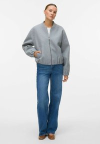 Femme avec les cheveux plaqués en arrière, portant une veste zippée grise clair, un jean bleu à jambes larges et des chaussures marron, se tenant avec une main dans la poche.