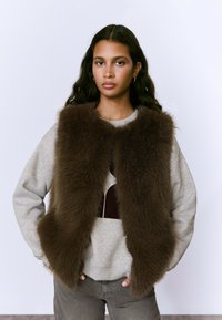 Chaleco de piel marrón sobre un sudadera gris, con un cuello redondo y textura suave. El fondo es de un color claro y uniforme.
