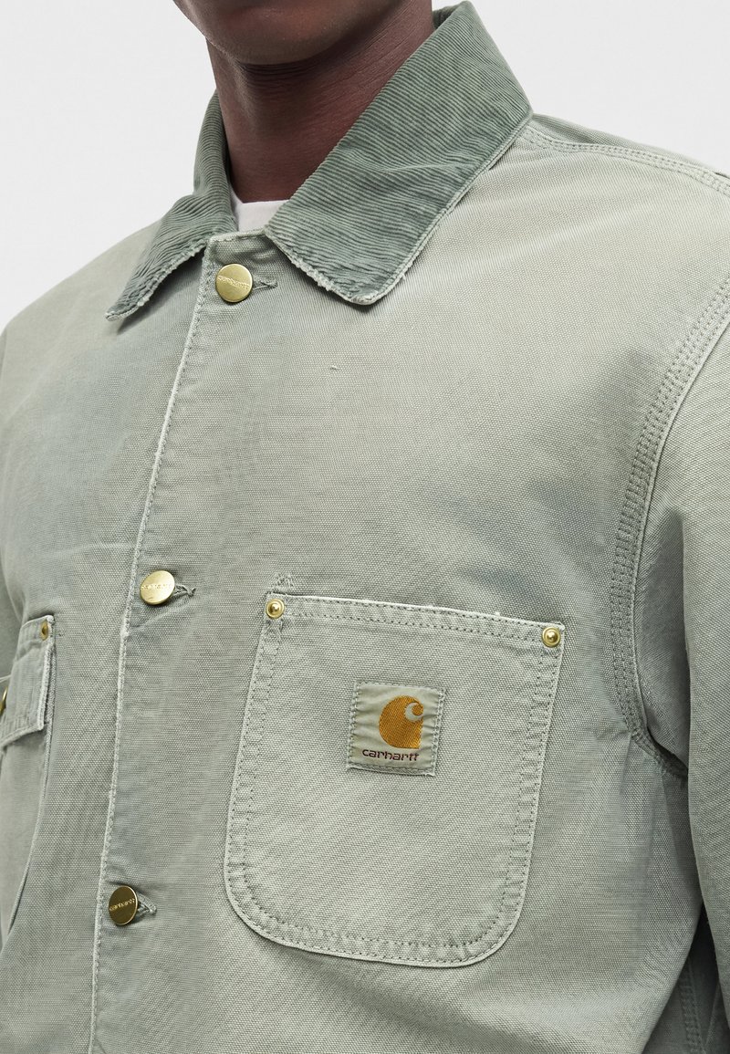 Mann trägt hellgrüne Carhartt-Jacke mit Cordkragen und goldenen Druckknöpfen, Nahaufnahme vom Brust- und Schulterbereich.