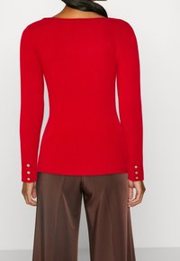 Geripptes rotes Langarmshirt mit weitem Ausschnitt und Akzenten aus drei Knöpfen an den Bündchen, kombiniert mit braunen Satinhosen mit weitem Bein.