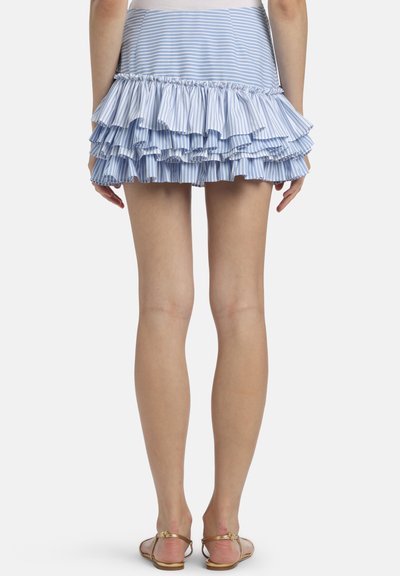 Person iført en blå- og hvidstribet mini-nederdel med flæser og gyldne flade sandaler, vist bagfra, stående mod en ensfarvet baggrund.