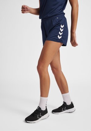 Personne portant un short de sport bleu marine avec des bandes blanches en chevron, des baskets noires et des chaussettes blanches, debout devant un fond blanc uni.