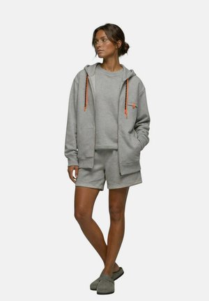 Femme portant un sweat à capuche gris avec des cordons orange, un short gris assorti, un pull et des chaussures grises sans lacets, debout avec une jambe croisée.