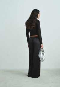 Schwarzes Langarmoberteil und weite schwarze Hose. Das Outfit wird von einer silbernen Metallic-Tasche mit einer zerknitterten Textur ergänzt. Rückansicht gezeigt.